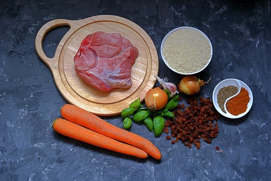 Ingredients for cooking pilaf on a dark gray concrete background. Meat on a wooden cutting board, carrots, rice, raisins, onion, garlic, spices.