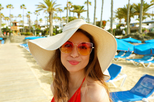 Close-up of attractive girl with long hair standing on the beach. Girl smiling to camera and shows her cool look. Straw hat on head. Palms trees on the background. Lanzarote, Canary Islands, Spain.
