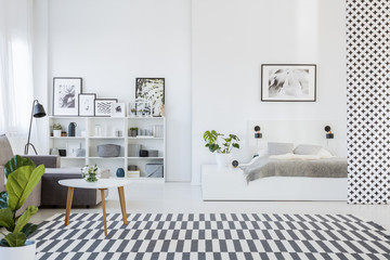 Real photo of an open space apartment interior with a cozy gray bed on a white platform and a wooden table next to a sofa