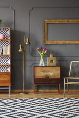 Gold lamp standing next to wooden cupboard with decor and fresh flowers in dark grey room interior with black and white carpet