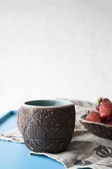 Clay cup with coffee or tea and a plate of strawberries in the background..