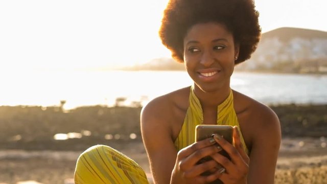 nice black race african young girl woman outdoor with smart phone checking email and social networks and connected with far friends during a nice sunset at the beach. beautiful people connect concept