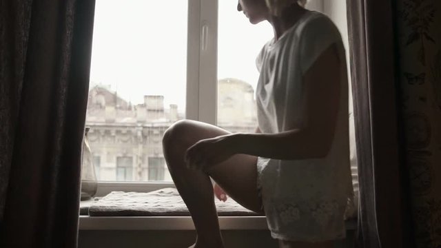 Young woman drinking coffee and looking through window while sitting at windowsill at home