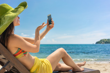 Attractive young woman sending online a love message through a selfie photo on the mobile phone while sitting on the beach during summer vacation