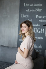 Pregnant blonde woman sits on a gray sofa in a beige dress in the interior of a loft