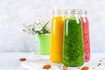 Green, yellow, purple smoothies in currant bottles, parsley, apple, kiwi, orange on a gray table with white flowers.