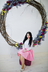Young brunette girl in pink skirt and white blouse posed indoor against large decorated wreath.