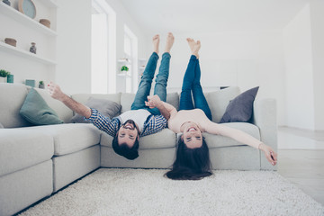 Portrait of childish, foolish couple in casual outfit jeans lying on sofa head over heels holding hands looking at camera in house white livingroom