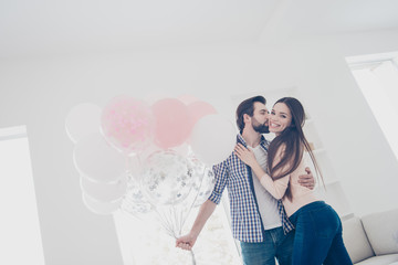 Low angle view portrait of attractive positive couple in casual outfits man preparing gift, present for  lover embracing kissing in cheek, standing in modern white apartment