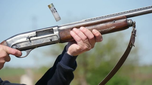 Closeup of a man keeping his single barrel rifle, aiming and shooting at clay targets. A spent cartidge falls down on a range in slow motion