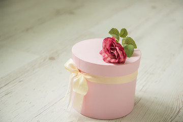 Pink round box with ribbon and rose on a light wooden background.
