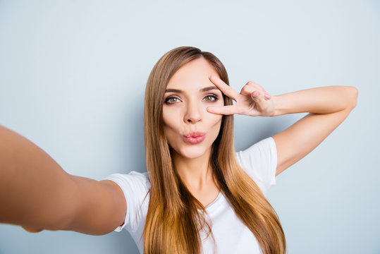 Self portrait of pretty cheerful girl shooting selfie on front camera gesturing v-sign near eye blowing kiss with pout lips isolated on grey background