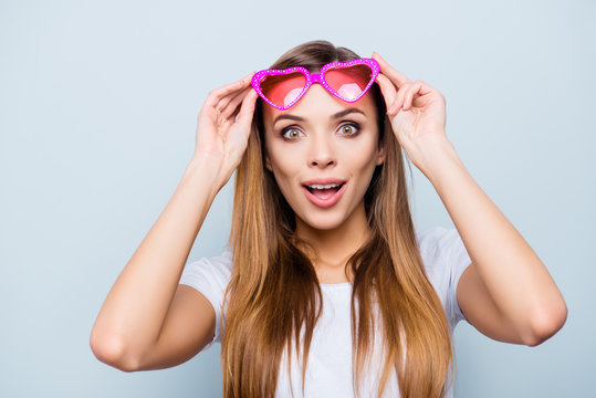 Portrait with copy space, empty place of impressed wondered girl raised her pink glasses in heart shape with wide open eyes mouth isolated on grey background
