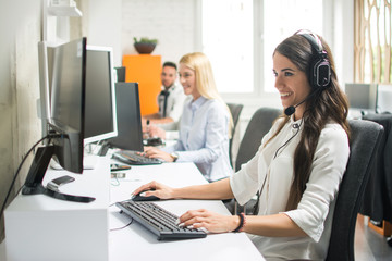 Portrait of beautiful and cheerful young woman telephone operator with headset working on desktop computer in row in customer service call support helpline business center with co-workers
