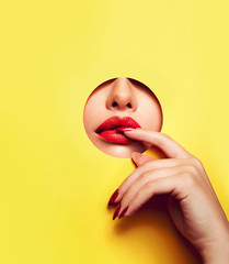 Red lips peep through the hole in the yellow paper. Fingers at the mouth with bright red...