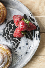 Delicious buns are sprinkled with sugar powder and red juicy strawberries lie on a light hand-made plate on a light wooden background.