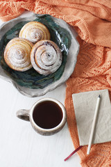 Three delicious beans sprinkled with sugar powder are placed on a hand-made plate, a cup of tea is standing next to it, a notepad and a pencil is lying. There is also an orange bright knitted plaid.