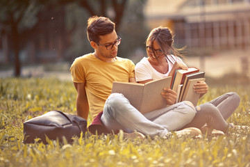 Two young students sitting at the campus yard , reading book and preparing for university exam.