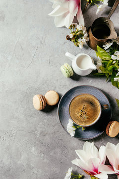 Blue cup of black espresso coffee french dessert macaroons, cream and spring flowers magnolia, blooming cherry branches over grey texture background. Top view, space. Spring greeting card