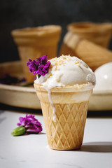 Homemade vanilla ice cream in small waffle cup served with purple edible flowers and metal spoon in ceramic plate on white marble kitchen table.