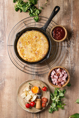 Spanish potato omelette tortilla with bacon served in cast-iron pan with sauce and ingredients above over wooden background. Top view, space.
