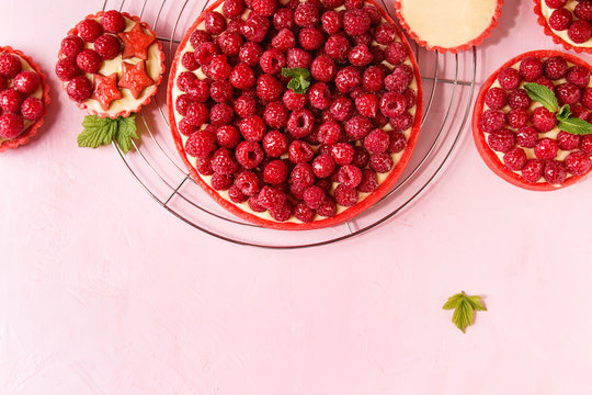 Variety of red raspberry shortbread tarts and tartlets with lemon custard and glazed fresh raspberries served on cooling rack over pink pastel background. Top view, copy space.