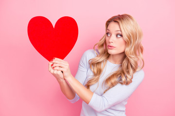 Portrait of doubting confused girl having love symbol in hands looking at big red carton heart figure isolated on pink background