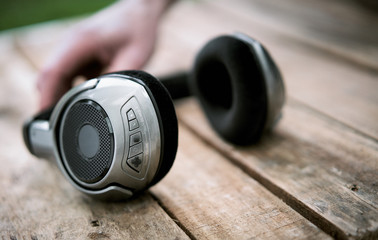 A 45 degree view of a set of wireless headphones and mans hand lying on a rustic wooden table outside. Styling and grain effect added to image.