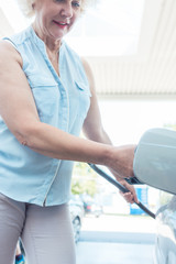 Fototapeta premium Low-angle view portrait of an active senior woman smiling while filling up the gas tank of her car at the station in summer