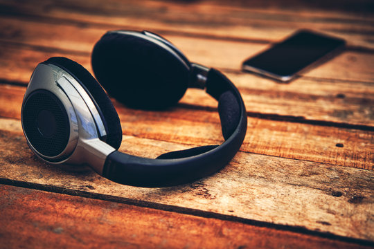 A 45 Degree View Of A Set Of Wireless Headphones And Mobile Phone Lying On A Rustic Wooden Table Outside. Styling And Grain Effect Added To Image.