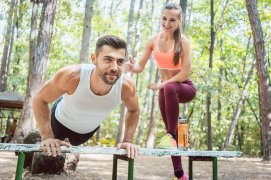 Sporty man doing push-up in an outdoor gym, his girlfriend is watching him 