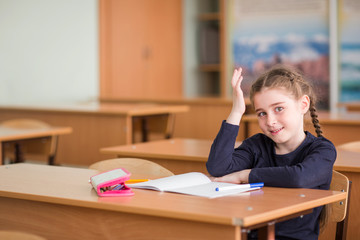 child girl sits at the Desk in the classroom, raises his hand and stares in surprise