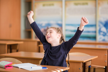 Happy child sits at the Desk in the classroom and shouts with joy with his hands up