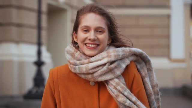 Portrait of content young woman wearing coat and scarf laughing to you with beautiful smile, while walking through historic centre of city on windy weather. Film look
