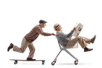Senior riding a longboard and pushing a shopping cart with another senior riding inside it and holding a newspaper © Ljupco Smokovski