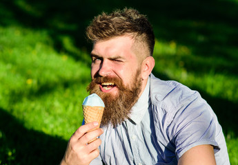 Temptation concept. Bearded man with ice cream cone. Man with long beard eats ice cream, while sits on grass. Man with beard and mustache on winking face enjoy ice cream, grass on background defocused