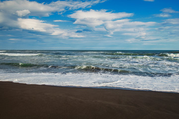Fototapeta na wymiar Halaktyr beach. Kamchatka. Russian federation. Dark almost black color sand beach of Pacific ocean. Stone mountains and yellow grass are on a background. Light blue sky