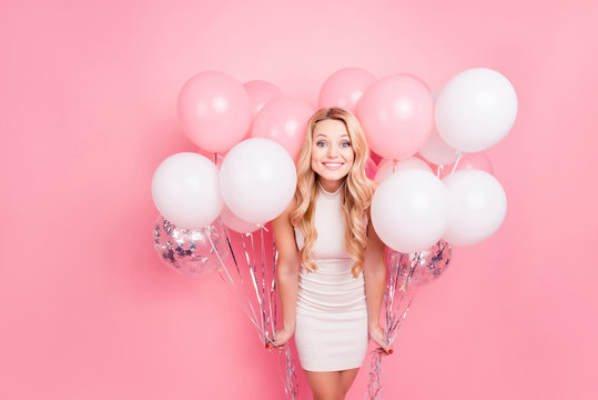 Portrait of positive elegant girlfriend in white dress having many white air balloons around her looking at camera isolated on pink background