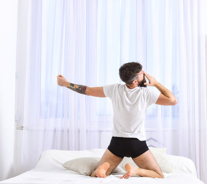 Guy yawning and stretching arm, full of energy in morning. Good morning concept. Man in shirt sits on bed, white curtains on background, rear view. Macho with beard stretching, relaxing after nap.