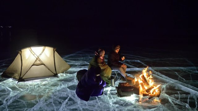 Three travelers by fire right on ice at night. Campground on ice. Tent stands next to fire. Lake Baikal. Nearby there is car. People are warming around campfire and are dressed in sleeping bags. This