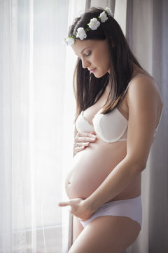 Pregnant happy woman in underwear by the window with curtains and a wreath of flowers on her head