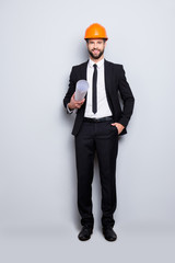 Full length size portrait of attractive cheerful joyful investor in orange hard hat and black tux, tuxedo with tie, looking at camera, isolated on grey background, having paper roll in hand