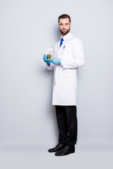 Full size fullbody portrait of attractive stylish scientist in white lab coat, black pants, shoes, tie holding test tubes with multi-colored liquid, looking at camera, isolated on grey background