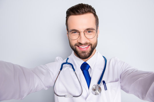Self portrait of joyful cheerful doc in white outfit with tie and stubble having stethoscope on his neck shooting selfie with two arms, isolated over grey background