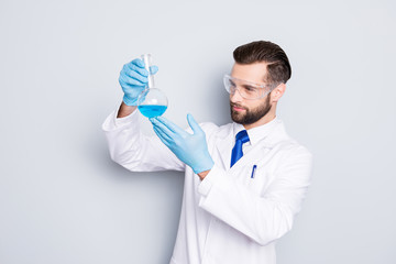 Portrait of concentrated busy scientist with stubble in white lab coat, gloves examine, looking at  flask with multi-colored liquid in his arm, isolated on grey background