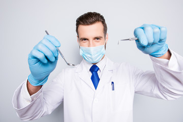 Naklejka premium Portrait of attractive hjandsome dentist in white lab coat, blue tie in working process on grey background, treat, examine cavity, searching for sick tooth, holding tools in hands