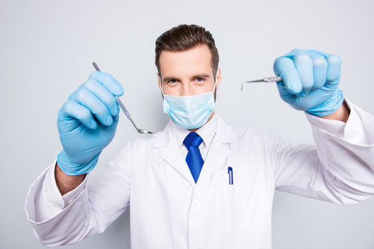 Portrait of attractive hjandsome dentist in white lab coat, blue tie in working process on grey background, treat, examine cavity, searching for sick tooth, holding tools in hands