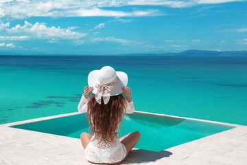 Fototapeta na wymiar Carefree Woman relaxing in infinity swimming pool looking at view. Luxury resort. Beautiful destination summer vactions. Back view of traveller girl in beach hat and white bikini relax on Maldives.