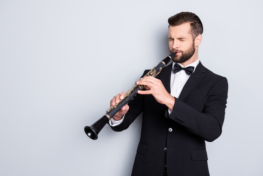 Portrait with copy space, empty place of stylish talented man with hairstyle in black tux with bow playing from heart on bassoon with closed eyes, isolated on grey background