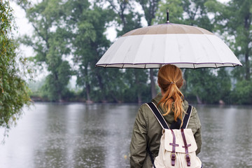 Woman with umbrella and backpack standing in rain on the shore of lake